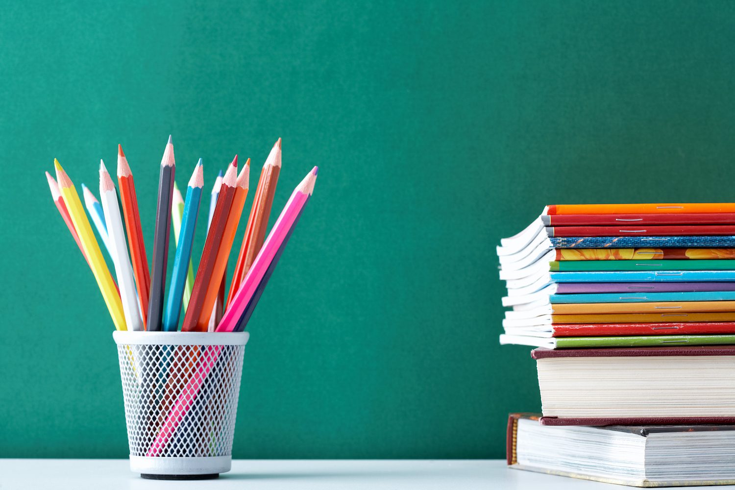 Image of colored pencils in a cup and a stack of books against a green chalkboard.
